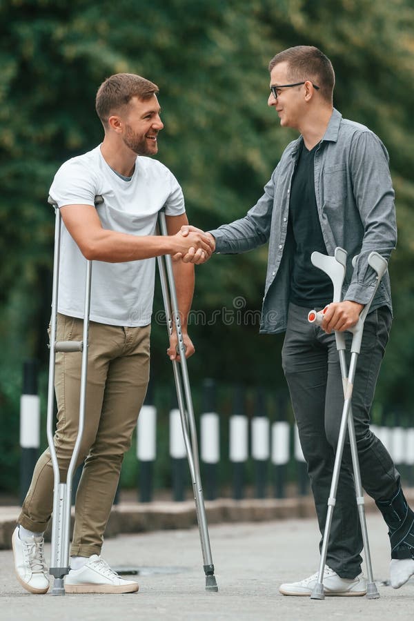 Doing Handshake. Two Men with Crutches is Outdoors on the Road Stock