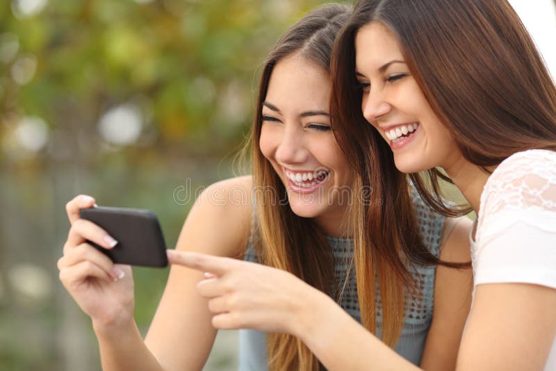 Dois amigos engraçados das mulheres que riem e que compartilham de meios em um telefone esperto foto de stock Dois amigos engraçados das mulheres que riem e que compartilham de meios em um telefone esperto foto de stock