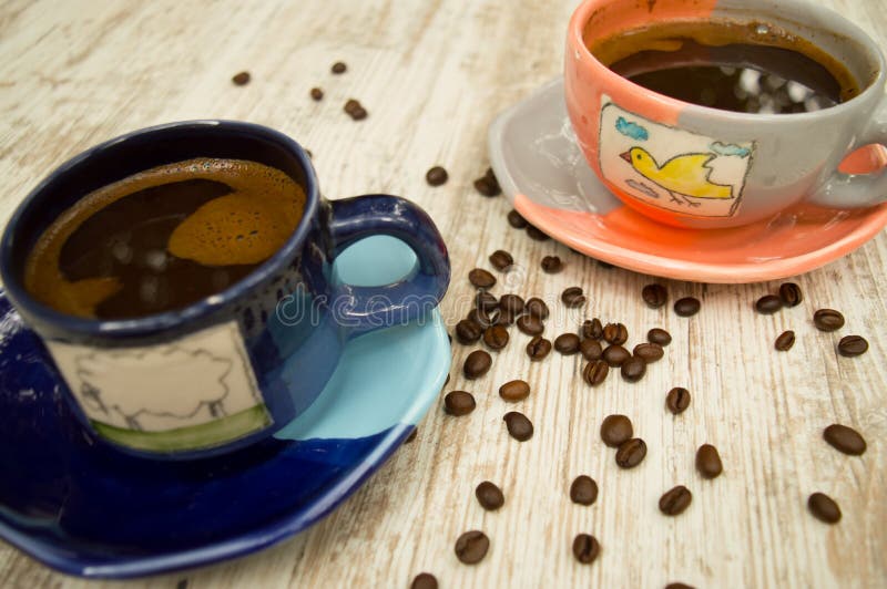 Two colorful coffee cups with black coffee and coffee yield scattered around. Black coffee. Two colorful coffee cups with black coffee and coffee yield scattered around. Black coffee.