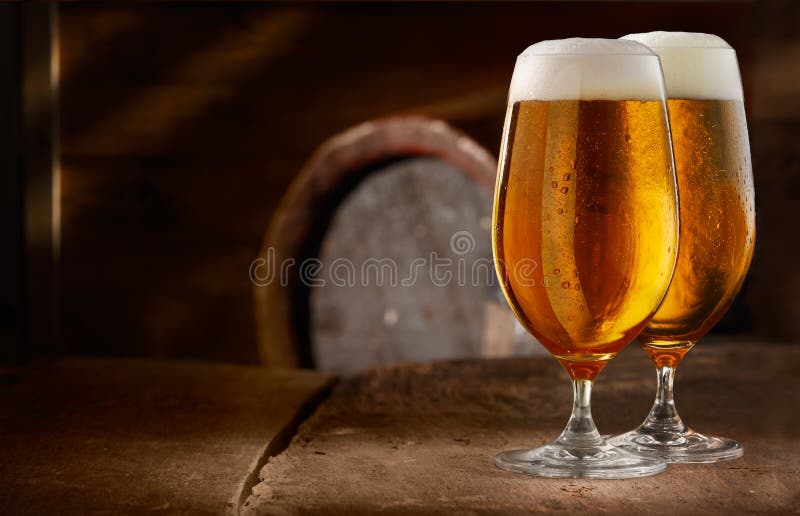 Two glasses of fresh foamy beer on a table in a vintage beer cellar with a barrel in the background. Two glasses of fresh foamy beer on a table in a vintage beer cellar with a barrel in the background