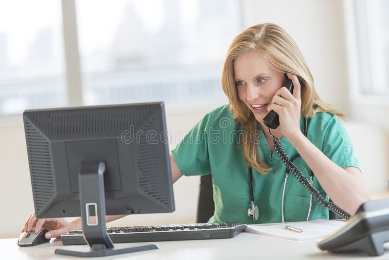 Doktor Using Computer While, das am Überlandleitungs-Telefon am Schreibtisch sich unterhält stockbild Doktor Using Computer While, das am Überlandleitungs-Telefon am Schreibtisch sich unterhält stockbild