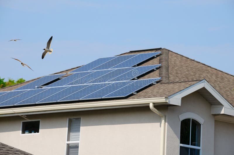 Home exterior showing house roof with rows of electric solar panels with the metaphor of green living being additionally portrayed with the flying birds. Home exterior showing house roof with rows of electric solar panels with the metaphor of green living being additionally portrayed with the flying birds