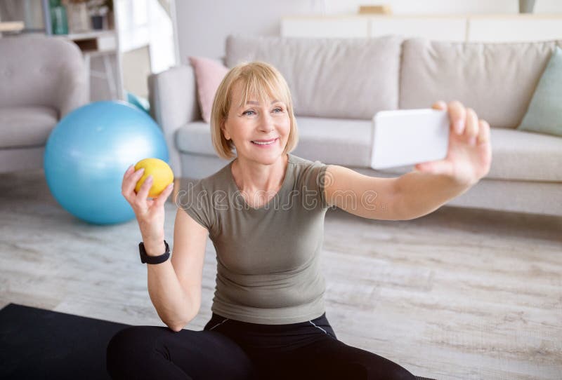 Domestic sports and healthy diet concept. Cheerful mature woman taking selfie with apple after home workout, indoors