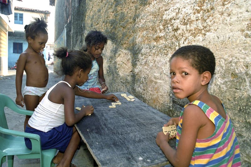 Dominó del juego de niños del Latino en los tugurios, Recife, el Brasil. El Brasil, Pernambuco, ciudad de Recife: escena de la calle de los tugurios Coelhos En el medio de la calle que juega muchachas y dominós de un juego del muchacho Una muchacha pone un pedazo del dominó y las tejas del dominó del control del muchacho en su mano Este juego de sociedad es también popular entre la juventud en América latina Una niña se coloca con un pezón en la boca para mirar el juego