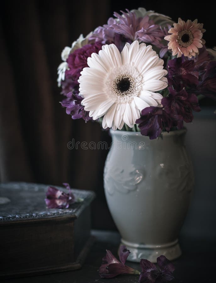 Still life image of flowers in a vase with muted colors and dark out of focus background. The flowers include a large white gerbera daisy and others with various shades of purple, magenta and peach. Still life image of flowers in a vase with muted colors and dark out of focus background. The flowers include a large white gerbera daisy and others with various shades of purple, magenta and peach.