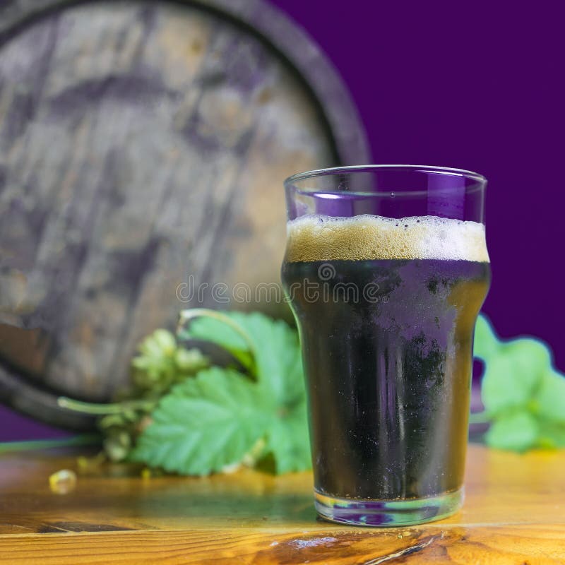 Donker bierglas op de lijst met hop op de achtergrond van vaten stock afbeelding Donker bierglas op de lijst met hop op de achtergrond van vaten stock afbeelding
