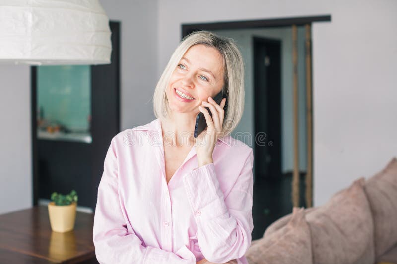 Donna adulta sorridente seduta su divano confortevole in soggiorno con cellulare fotografia stock