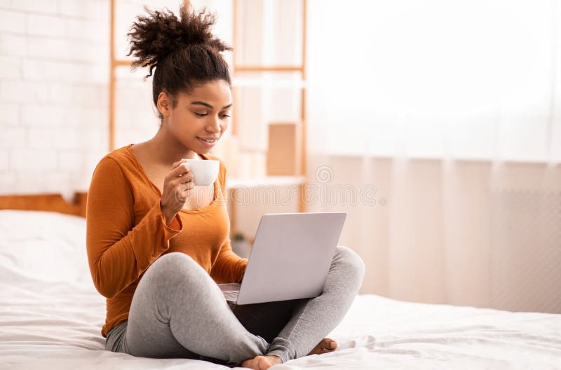 African American Lady Using Laptop Having Coffee Sitting On Bed At Home Relaxing On Weekend Morning. Empty Space, Selective Focus. African American Lady Using Laptop Having Coffee Sitting On Bed At Home Relaxing On Weekend Morning. Empty Space, Selective Focus
