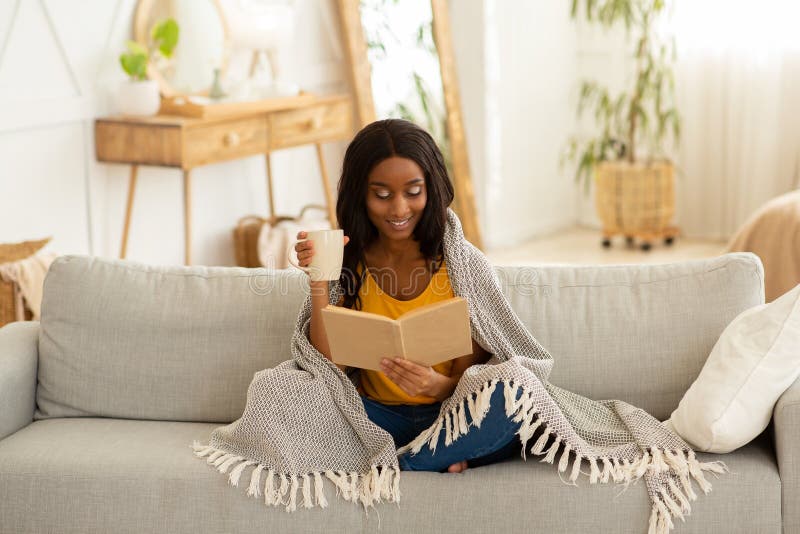 Beautiful African American lady with cup of fresh hot coffee reading fascinating book while cuddling under warm plaid on couch at home. Lazy weekend pastimes concept. Beautiful African American lady with cup of fresh hot coffee reading fascinating book while cuddling under warm plaid on couch at home. Lazy weekend pastimes concept