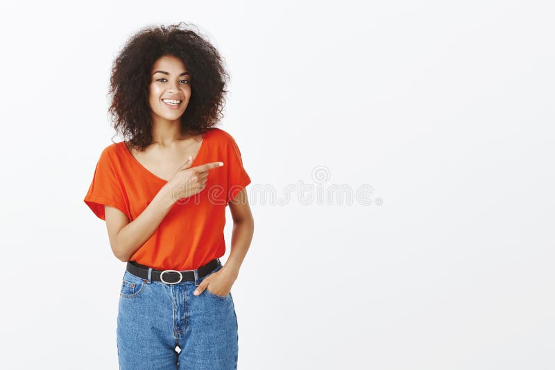 Donna afroamericana spensierata felice con l'acconciatura riccia che sorride largamente mentre indicando a sinistra e stando cont fotografie stock libere da diritti Donna afroamericana spensierata felice con l'acconciatura riccia che sorride largamente mentre indicando a sinistra e stando cont fotografie stock libere da diritti