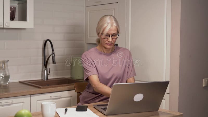 Donna anziana che digita laptop in cucina, riflettendo la concentrazione e la concentrazione archivi video