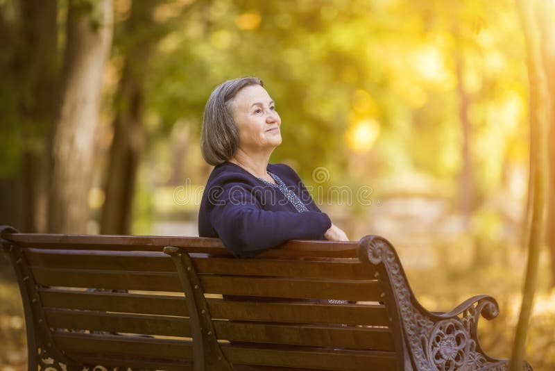 Donna anziana che si siede su un banco nel parco di autunno immagine stock Donna anziana che si siede su un banco nel parco di autunno immagine stock