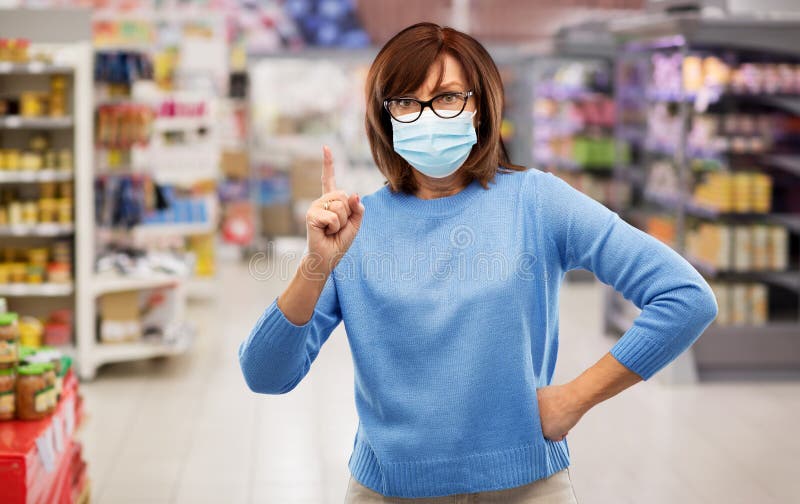 Health, safety and pandemic concept - angry senior woman in glasses wearing protective medical mask for protection from virus and pointing finger up over supermarket background. Health, safety and pandemic concept - angry senior woman in glasses wearing protective medical mask for protection from virus and pointing finger up over supermarket background