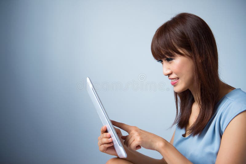 Portrait of Asian woman reading a tablet pc indoor wearing blue dress isolated on blue background. Asian female model. Portrait of Asian woman reading a tablet pc indoor wearing blue dress isolated on blue background. Asian female model.