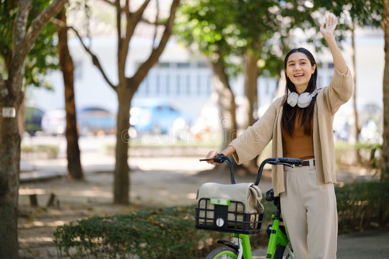 Donna asiatica felice che va in bicicletta in un parco pubblico e che saluta la mano. immagine stock libera da diritti