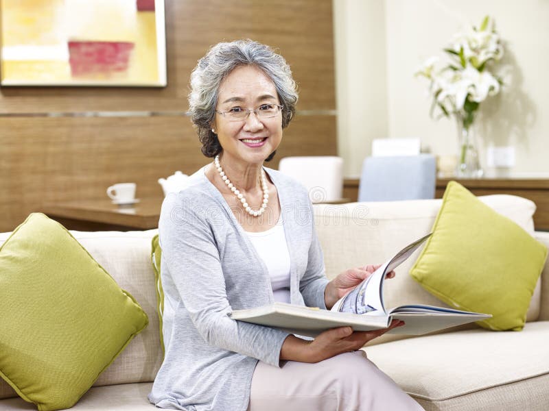 Senior asian woman sitting on couch at home holding a book look at camera smiling. Senior asian woman sitting on couch at home holding a book look at camera smiling.