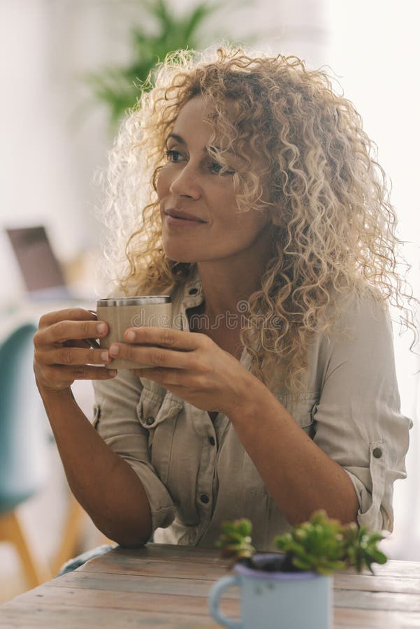 Donna attraente con lunghi capelli biondi ricci che si gode il tempo in casa da sola a bere un caffè o un tè e a guardare immagini stock