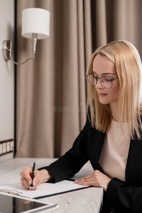 Donna bionda attraente in vetri e un funzionamento del vestito su una compressa, parlante sul telefono e prendente le note in a fotografie stock Donna bionda attraente in vetri e un funzionamento del vestito su una compressa, parlante sul telefono e prendente le note in a fotografie stock
