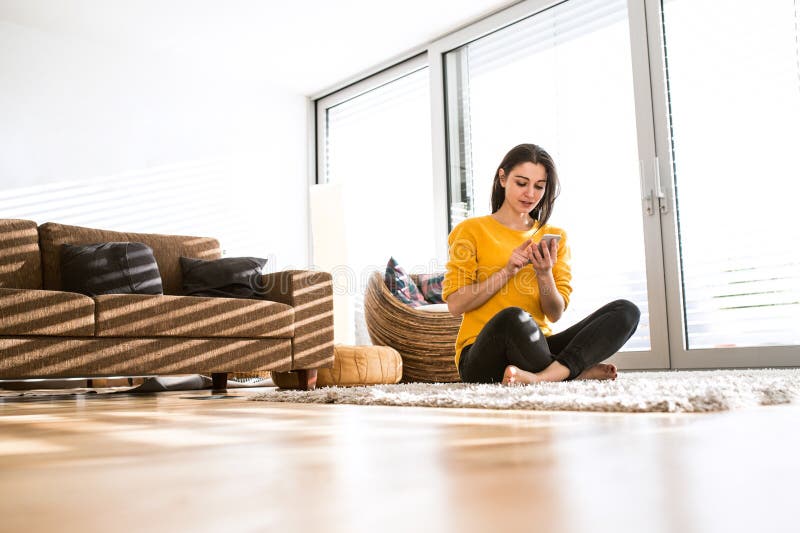 Donna a casa che si siede sul tappeto, tenendo Smart Phone, mandante un sms fotografie stock libere da diritti Donna a casa che si siede sul tappeto, tenendo Smart Phone, mandante un sms fotografie stock libere da diritti