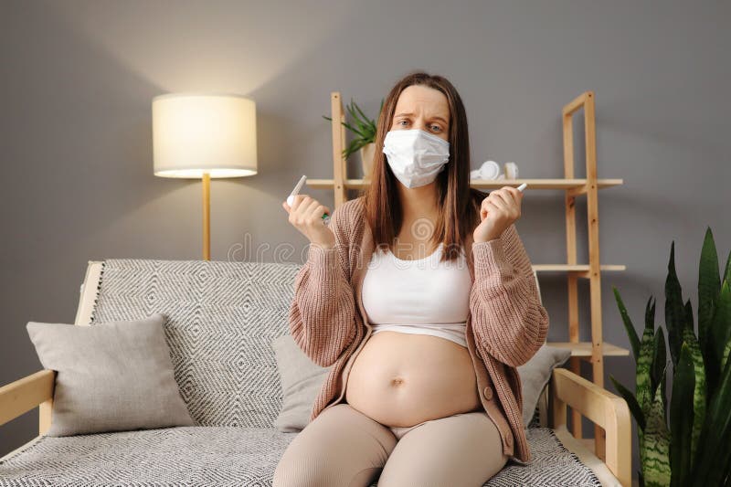 Sick ill unhealthy Caucasian pregnant woman sitting on sofa at home holding nasal spray and spray for inhalation taking care of her health during pregnancy flu. Sick ill unhealthy Caucasian pregnant woman sitting on sofa at home holding nasal spray and spray for inhalation taking care of her health during pregnancy flu