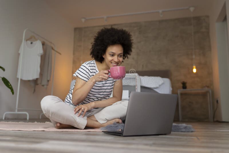 Beautiful young mixed race woman sitting on the bedroom floor, having a video call on a laptop computer and drinking coffee. Beautiful young mixed race woman sitting on the bedroom floor, having a video call on a laptop computer and drinking coffee