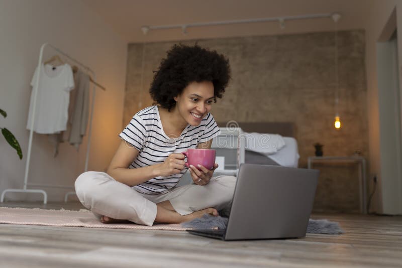 Beautiful young mixed race woman sitting on the bedroom floor, having a video call on a laptop computer and drinking coffee. Beautiful young mixed race woman sitting on the bedroom floor, having a video call on a laptop computer and drinking coffee