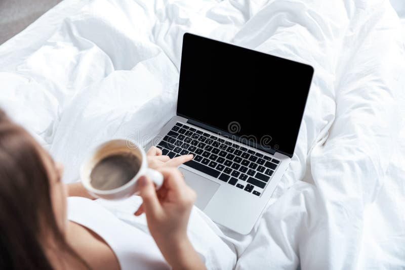 Donna che lavora al computer portatile e che mangia il caffè di mattina a letto fotografia stock Donna che lavora al computer portatile e che mangia il caffè di mattina a letto fotografia stock