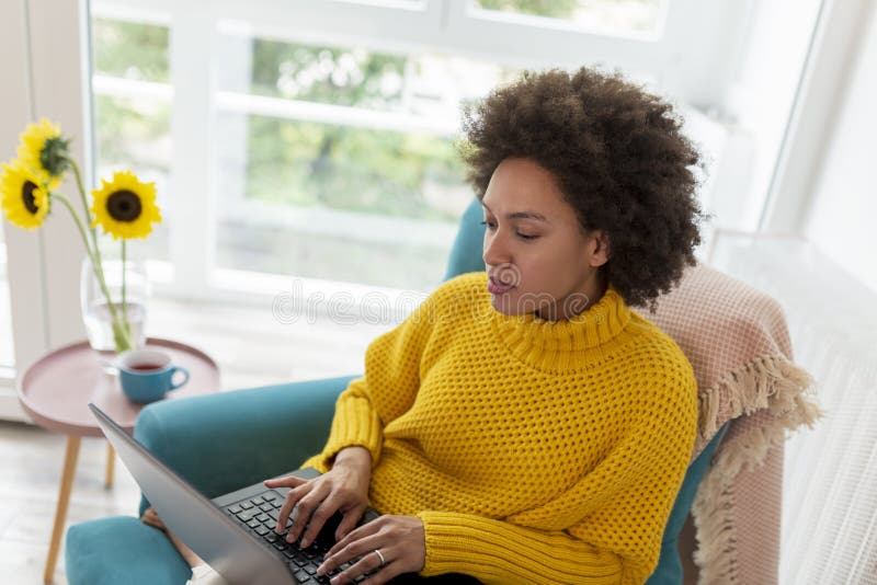 Beautiful young mixed race woman sitting in an armchair, working remotely from home, using a laptop computer. Beautiful young mixed race woman sitting in an armchair, working remotely from home, using a laptop computer