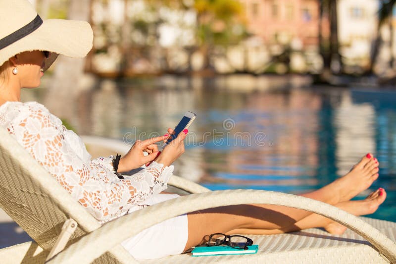 Donna che si siede nella sedia dalla piscina e che per mezzo dello smartphone fotografia stock Donna che si siede nella sedia dalla piscina e che per mezzo dello smartphone fotografia stock