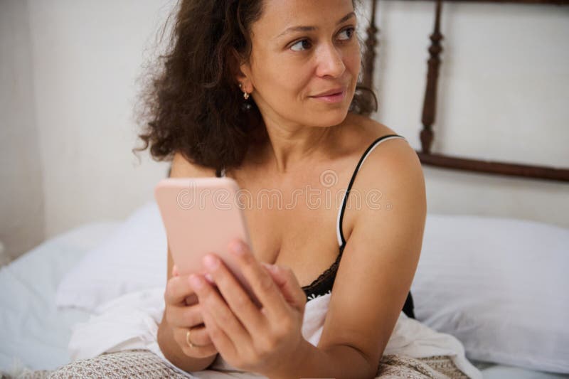 Donna che tiene in mano uno smartphone mentre è seduta su un letto in condizioni di illuminazione variabile. Una donna si siede comodamente a letto tenendo il telefono con un'espressione pensierosa in una camera da letto con mobili di legno e illuminazione calma raffigurata in un momento di pacifica contemplazione al chiuso.