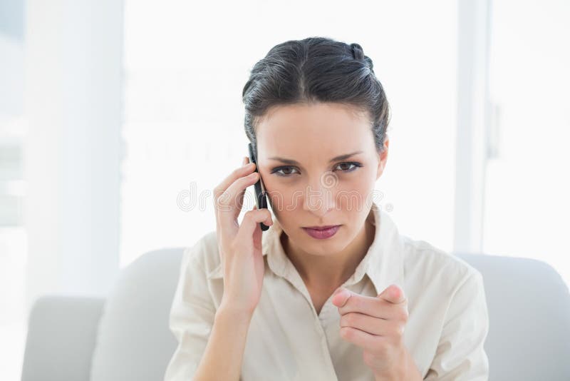 Donna di affari castana alla moda irritata che fa una telefonata e che indica macchina fotografica con il suo dito immagini stock Donna di affari castana alla moda irritata che fa una telefonata e che indica macchina fotografica con il suo dito immagini stock