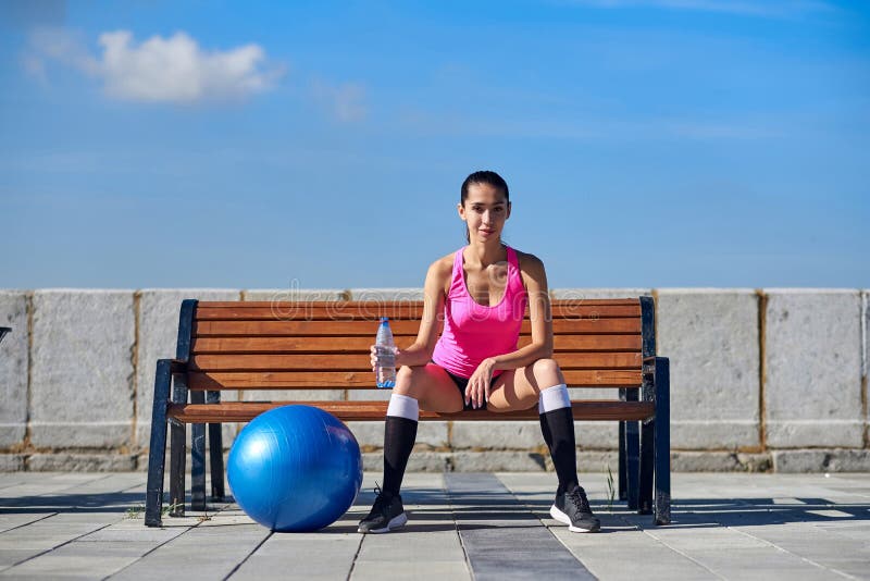 Donna di forma fisica con la bottiglia e la palla adatta di gomma Acqua potabile femminile dopo la formazione sul banco in città immagine stock Donna di forma fisica con la bottiglia e la palla adatta di gomma Acqua potabile femminile dopo la formazione sul banco in città immagine stock