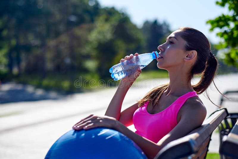 Donna di forma fisica con la bottiglia e la palla adatta di gomma Acqua potabile femminile dopo la formazione sul banco nel parco fotografia stock libera da diritti Donna di forma fisica con la bottiglia e la palla adatta di gomma Acqua potabile femminile dopo la formazione sul banco nel parco fotografia stock libera da diritti