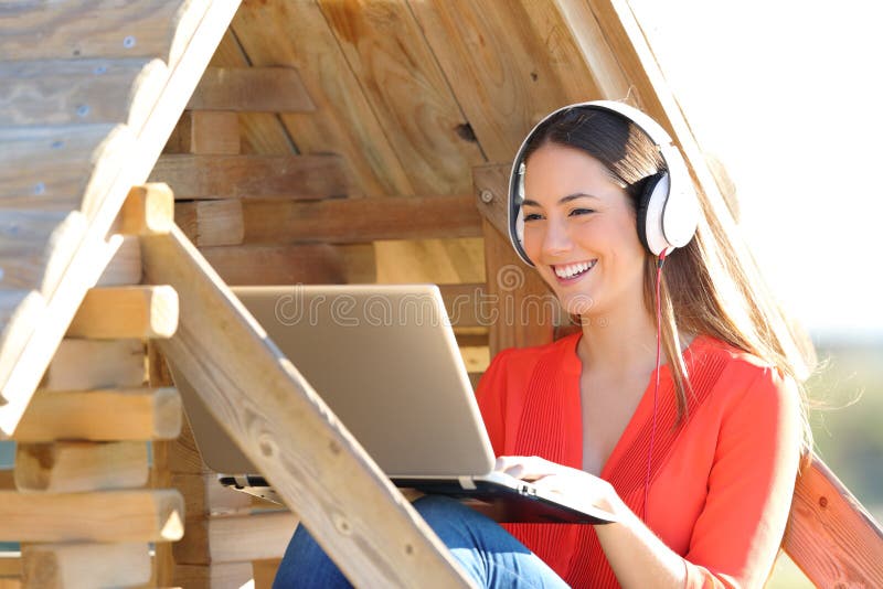 Donna felice che utilizza computer portatile e le cuffie in una casa di legno fotografia stock Donna felice che utilizza computer portatile e le cuffie in una casa di legno fotografia stock