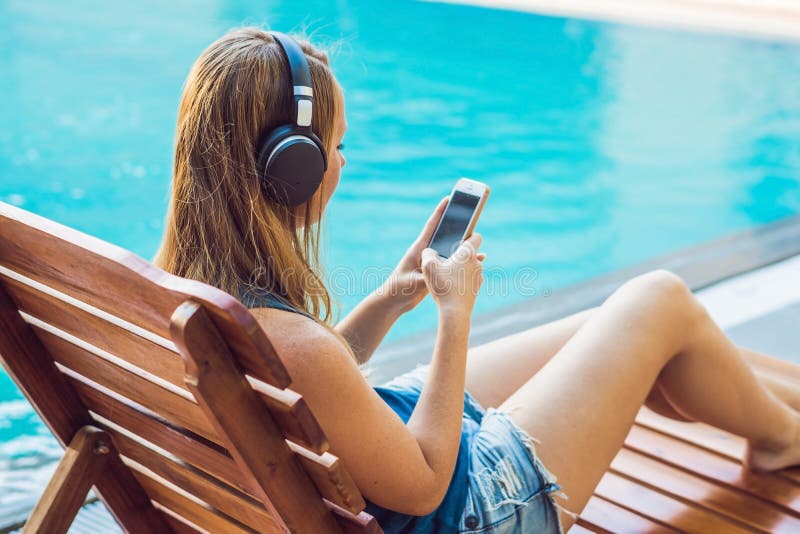 Happy smartphone woman relaxing near swimming pool listening with earbuds to streaming music. Beautiful girl using her mobile phone app 4g data to play songs while relaxing on summer luxury vacations. Happy smartphone woman relaxing near swimming pool listening with earbuds to streaming music. Beautiful girl using her mobile phone app 4g data to play songs while relaxing on summer luxury vacations.