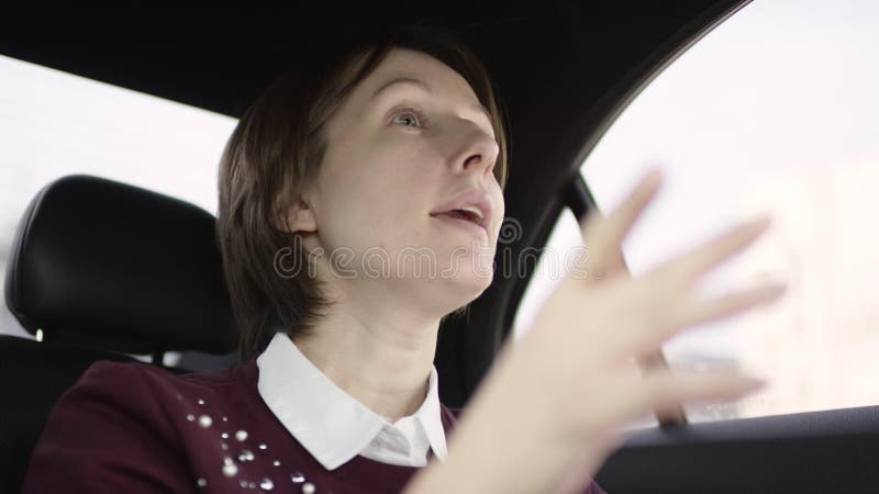 Donna graziosa con capelli marroni in blusa bianca e nella guida marrone rossiccio del maglione in automobile e spiegare il giust