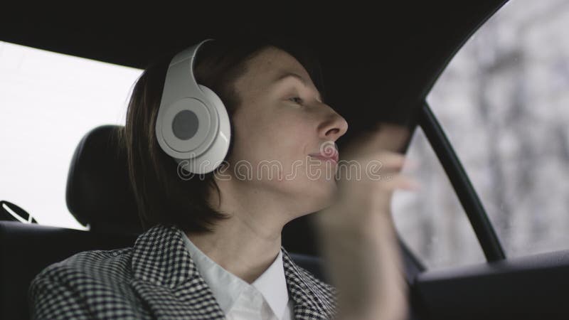 Donna graziosa con capelli marroni nella guida bianca del cappotto del controllo e della blusa in automobile ed ascoltare musica