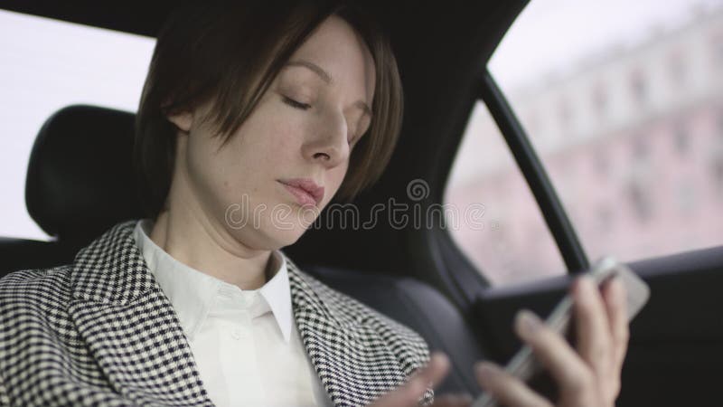 Donna graziosa con capelli marroni nella guida bianca del cappotto del controllo e della blusa in automobile e messaggio di batti