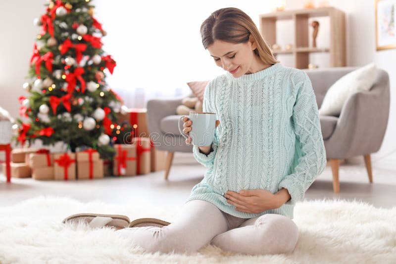Donna incinta con la tazza di tè che si siede sul pavimento nella sala decorata per il Natale fotografie stock Donna incinta con la tazza di tè che si siede sul pavimento nella sala decorata per il Natale fotografie stock