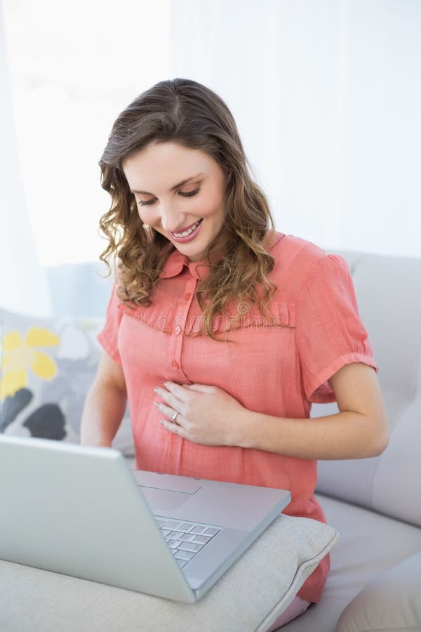 Donna incinta pacifica che lavora con il suo computer portatile che si siede su uno strato nel salone immagini stock Donna incinta pacifica che lavora con il suo computer portatile che si siede su uno strato nel salone immagini stock