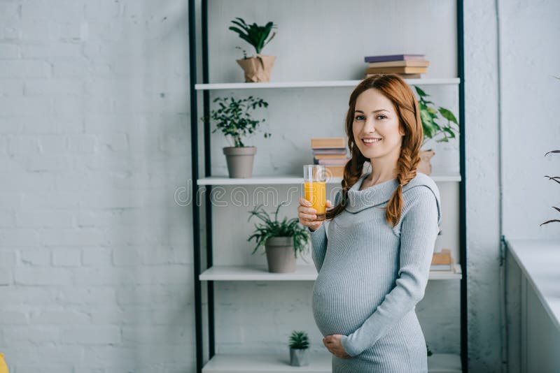 donna incinta sorridente attraente che tiene vetro di succo d'arancia e che esamina macchina fotografica fotografia stock libera da diritti donna incinta sorridente attraente che tiene vetro di succo d'arancia e che esamina macchina fotografica fotografia stock libera da diritti