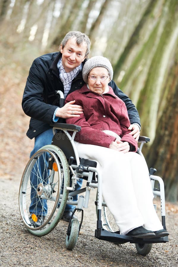 Caregiver careful men walking and embracing disabled senior women grandmother at wheelchair in nature. Caregiver careful men walking and embracing disabled senior women grandmother at wheelchair in nature