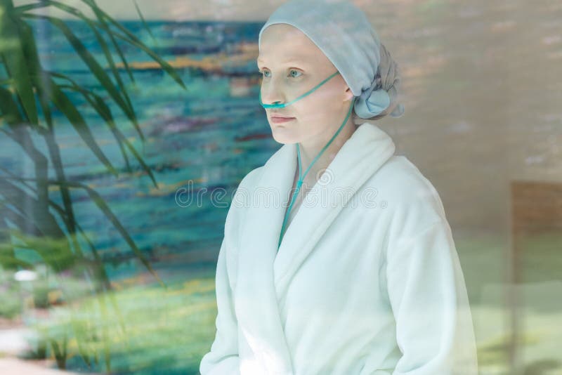 Sick woman in headscarf and nasal cannula looking through window. Sick woman in headscarf and nasal cannula looking through window