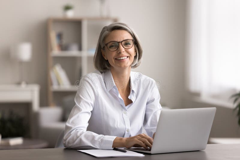 Donna media felice siede in ufficio con il computer portatile immagine stock