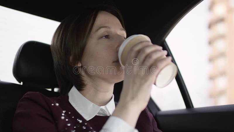 Donna piacevole con capelli marroni in blusa bianca e nella guida marrone rossiccio del maglione in automobile e bere il suo caff