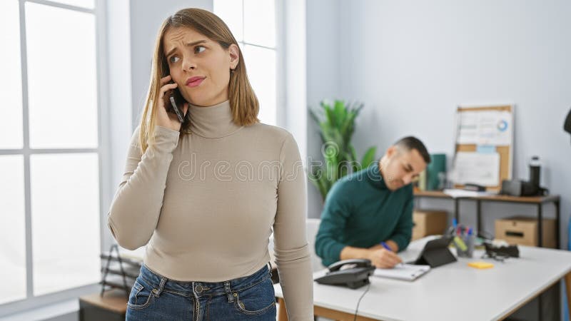 Donna preoccupata al telefono con un uomo concentrato che lavora dietro in un ufficio luminoso fotografia stock