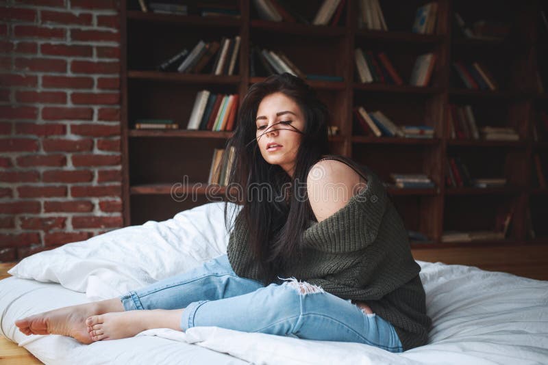 Donna sensuale con capelli scuri in maglione verde e jeans che si siedono su un letto immagini stock libere da diritti Donna sensuale con capelli scuri in maglione verde e jeans che si siedono su un letto immagini stock libere da diritti