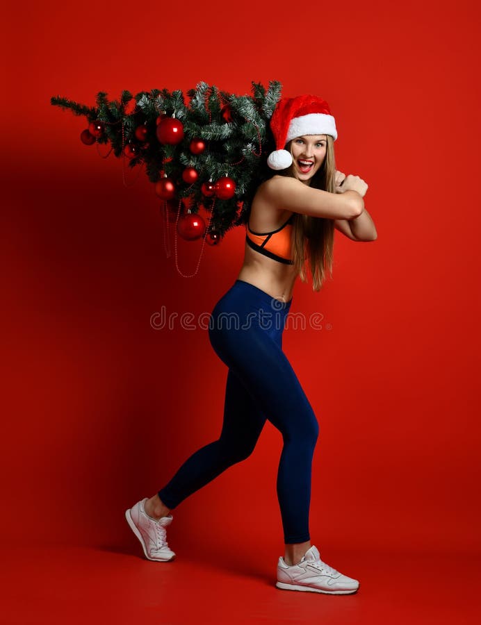 Donna sexy di sport di forma fisica di Natale che porta il cappello di Santa che tiene l'albero di natale sulle sue spalle fotografie stock Donna sexy di sport di forma fisica di Natale che porta il cappello di Santa che tiene l'albero di natale sulle sue spalle fotografie stock