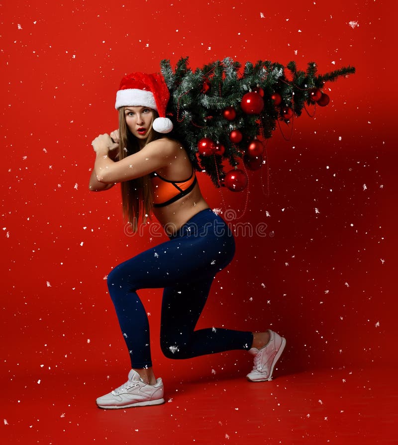 Donna sexy di sport di forma fisica di Natale che porta il cappello di Santa che tiene l'albero di natale sulle sue spalle Fiocch fotografia stock Donna sexy di sport di forma fisica di Natale che porta il cappello di Santa che tiene l'albero di natale sulle sue spalle Fiocch fotografia stock