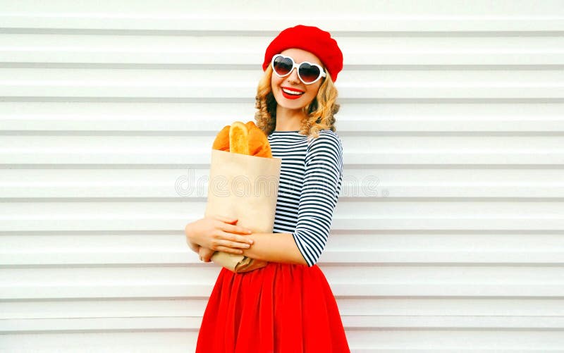 Donna sorridente affascinante del ritratto che porta berretto rosso francese che tiene sacco di carta con le baguette lunghe del immagini stock libere da diritti